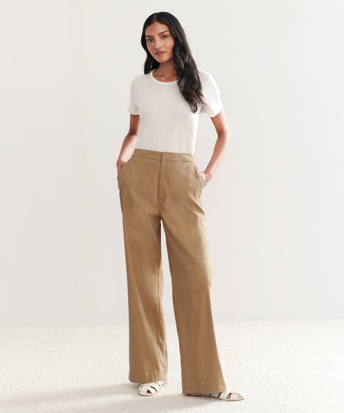 A woman stands against a neutral background wearing a white t-shirt, the high-waisted Linen Keaton Pant in khaki, and white sandals, with her hands in her pockets.