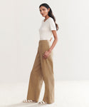 A woman with long, wavy dark hair and a neutral expression stands sideways in a minimal, light-colored room wearing a white short-sleeve top and the high-waisted khaki Linen Keaton Pant, styled with white flat sandals.