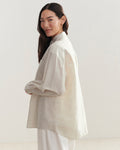 A woman with long dark hair, dressed in an Oversized Shirt and white pants, stands sideways against a light background, smiling softly over her shoulder.