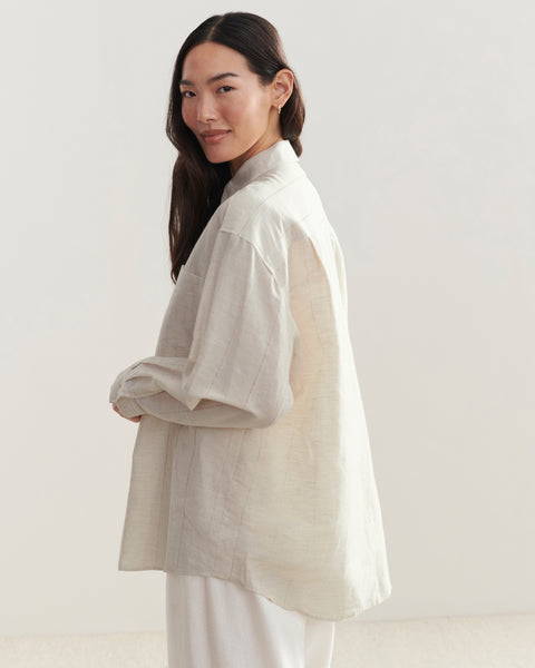 A woman with long dark hair, dressed in an Oversized Shirt and white pants, stands sideways against a light background, smiling softly over her shoulder.