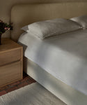 A neatly made bed with the Dusk Linen Duvet Cover and pillows sits beside a light wood nightstand on a textured white rug and brick floor, with a small vase of flowers—a picture of luxury bedding.