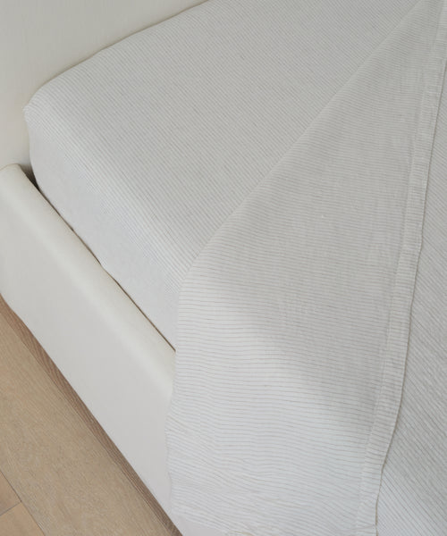 Close-up of a neatly made bed with the Dusk Linen Fitted Sheet in white featuring subtle vertical stripes, on a light-colored bed frame beside a wooden floor. Crafted from premium materials and made in Portugal.