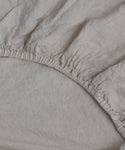 Close-up of the Dusk Linen Fitted Sheet with gathered elastic edges, partially stretched over a mattress. Lightweight and expertly crafted in Portugal for a relaxed look and perfect fit.