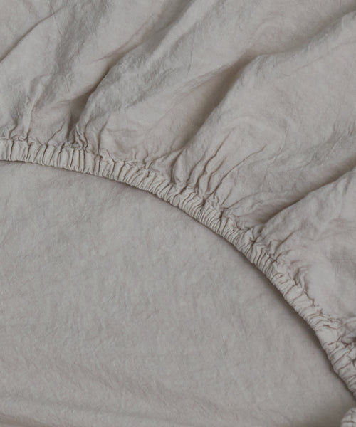 Close-up of the Dusk Linen Fitted Sheet with gathered elastic edges, partially stretched over a mattress. Lightweight and expertly crafted in Portugal for a relaxed look and perfect fit.