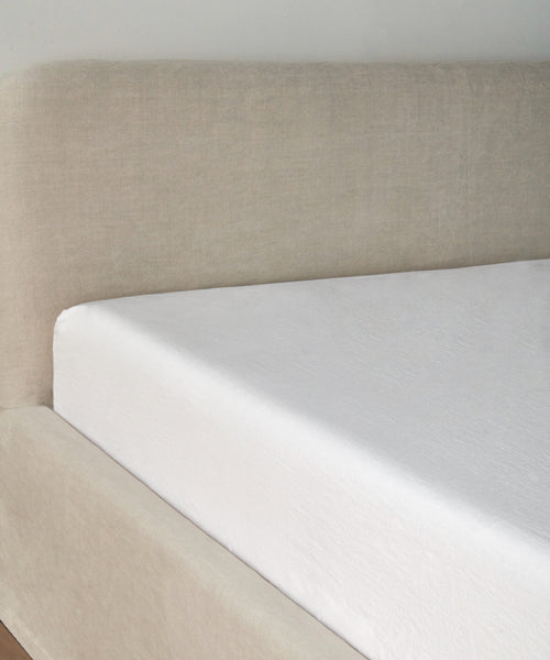 A close-up of a neatly made bed with a light beige upholstered headboard, showcasing the Dusk Linen Fitted Sheet—lightweight, comfortable, and made in Portugal—set against a plain light-colored wall.