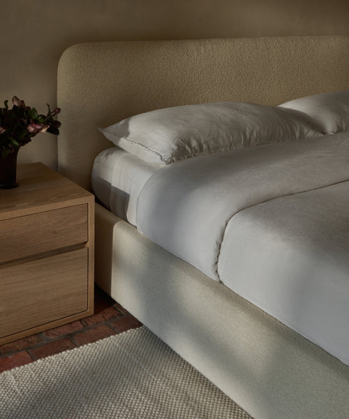 A neatly made bed with the Dusk Linen Fitted Sheet and pillows sits beside a wooden nightstand with a vase of dark flowers, on a light textured rug over a brick floor in a softly lit room.