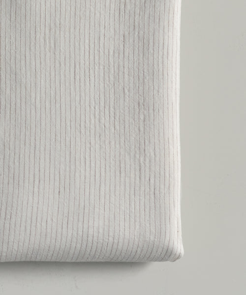 A close-up of the Dusk Linen Flat Sheet, folded in light beige with thin gray pinstripes. Its soft, smooth texture and subtle flax stripe pattern evoke timeless luxury bedding on a light surface.