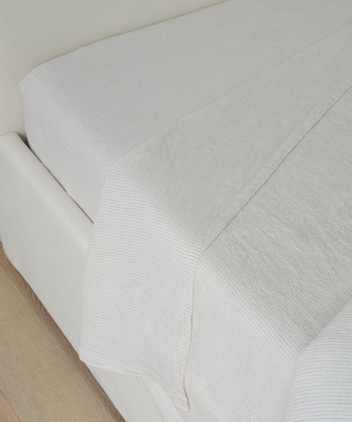A neatly made bed features the Dusk Linen Flat Sheet, boasting a subtle flax stripe texture, paired with a white fitted sheet on a light-colored frame beside a wooden floor.