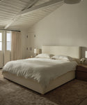 Minimalist bedroom with a large bed dressed in luxury bedding and a Dusk Linen Flat Sheet, two wooden nightstands with lamps, beige carpet, cream walls, curtained window, and exposed white ceiling beams filled with soft natural light.