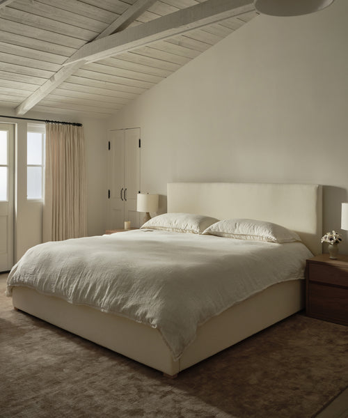 Minimalist bedroom with a large bed dressed in luxury bedding and a Dusk Linen Flat Sheet, two wooden nightstands with lamps, beige carpet, cream walls, curtained window, and exposed white ceiling beams filled with soft natural light.