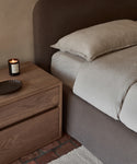 A neatly made bed featuring the Dusk Linen Flat Sheet in light beige with a brown upholstered headboard sits beside a wooden nightstand holding a lit candle and dark dish, atop a brick floor with a woven rug visible.