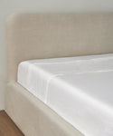 A close-up of a neatly made bed with a beige upholstered headboard and frame, dressed in crisp white Dusk Linen Flat Sheets on a smooth surface, set atop a wooden floor.