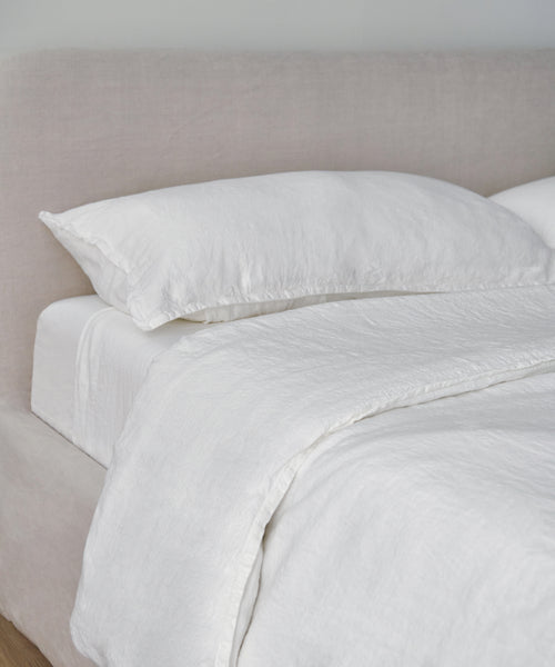 A neatly made bed with a light beige upholstered headboard, dressed in luxury bedding including the Dusk Linen Flat Sheet, pillow, and duvet, creates a clean and minimalistic look.