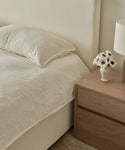 A neatly made bed with cream bedding is topped with the Dusk Linen Pillow Sham Set of 2 in lightweight linen, beside a wooden nightstand holding a vase of white flowers and a beige lamp—creating a calm, minimalist look.