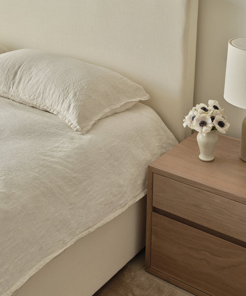 A neatly made bed with cream bedding is topped with the Dusk Linen Pillow Sham Set of 2 in lightweight linen, beside a wooden nightstand holding a vase of white flowers and a beige lamp—creating a calm, minimalist look.