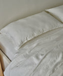 A neatly made bed features luxury white linens and a Dusk Linen Pillow Sham Set of 2, along with a comforter, all set against a light-colored wall.
