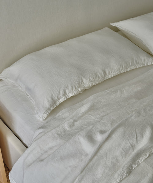 A neatly made bed features luxury white linens and a Dusk Linen Pillow Sham Set of 2, along with a comforter, all set against a light-colored wall.