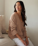 A woman with long dark hair sits on a beige couch, wearing the Linen Dylan Sweater and white pants, looking thoughtfully to the side in a softly lit, neutral-toned room.