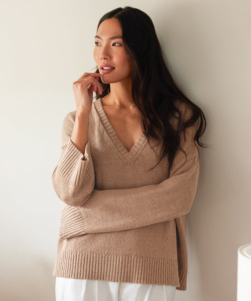 A woman with long dark hair, wearing the beige Linen Dylan Sweater and white pants, stands against a light-colored wall with her arms crossed, gazing thoughtfully to the side.