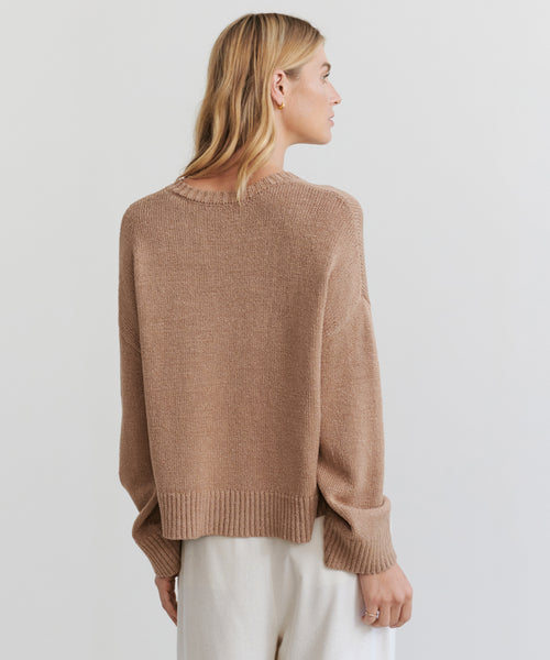 A blonde woman stands facing away against a plain light background, wearing the Linen Dylan Sweater in a loose, light brown summer knit style, paired with white pants.