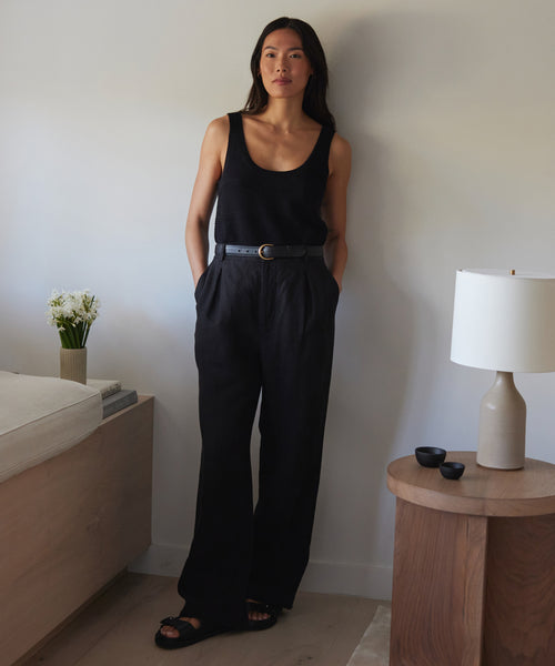 A woman stands indoors against a wall, wearing black Linen Kyle Trousers with a matching tank top, belt, and sandals. She has her hands in her pockets. A lamp, small bowls, and a vase of flowers are nearby.