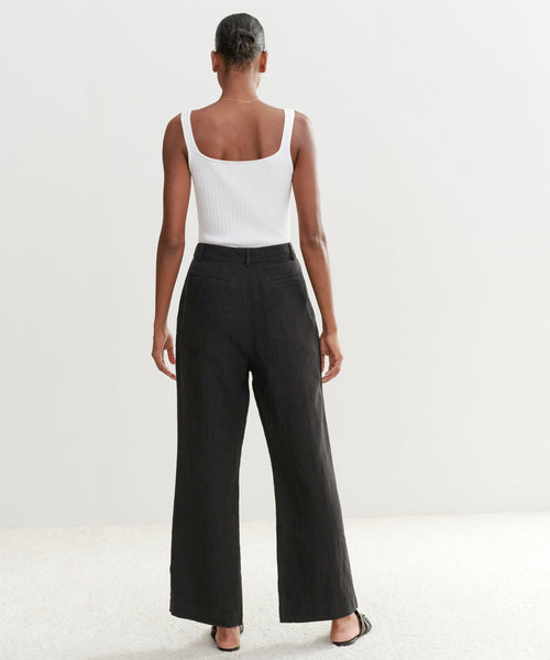 A person stands facing away in a white ribbed tank top, black Linen Kyle Trousers, and black sandals against a plain light background. Their hair is styled in a neat bun.