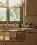 A rustic kitchen sink with a brass faucet and the Laurel Hand Towel in a soft linen cotton blend draped over the edge, alongside a bowl of artichokes and pears. Sunlight filters through sheer curtains, highlighting greenery outside.