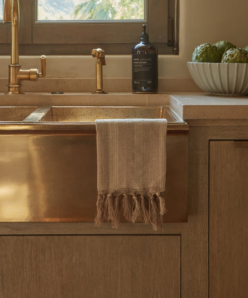 The beige Laurel Hand Towel, woven in Turkey from a linen-cotton blend, hangs over a brass kitchen sink with gold fixtures. Nearby, a bottle of soap and bowl of artichokes rest on the tan countertop by the window.