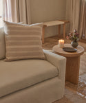 A beige sofa with the Laurel Pillow sits beside a wooden side table holding a lit candle, a small dish, and a vase of flowers. Neutral curtains and a bench appear in the background.