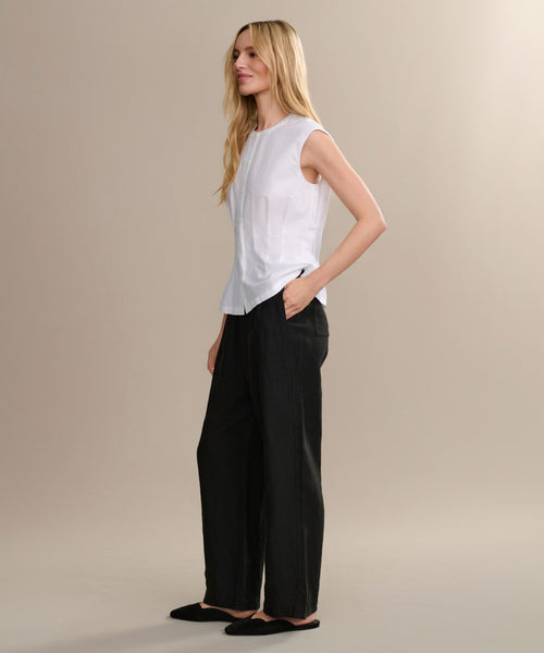 A woman with long blonde hair wears a sleeveless white top, black Linen Logan Pant, and black flats. She stands in profile with one hand in her pocket against a plain beige background—perfect for everyday style.
