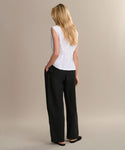 A woman with long blonde hair stands facing away in a sleeveless white top, black Linen Logan Pant, and black flats against a beige background—ideal for effortless everyday outfitting.