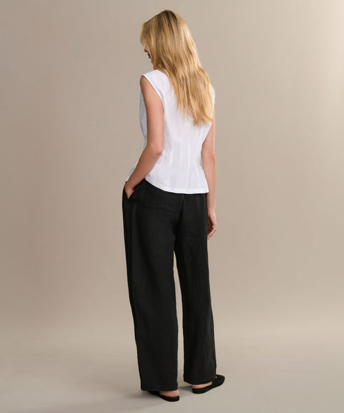 A woman with long blonde hair stands facing away in a sleeveless white top, black Linen Logan Pant, and black flats against a beige background—ideal for effortless everyday outfitting.