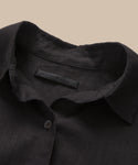 A close-up of the Linen Logan Shirt, a black boxy-fit button-up with a single button visible and an inner collar tag reading jenn kayne, set against a beige background.