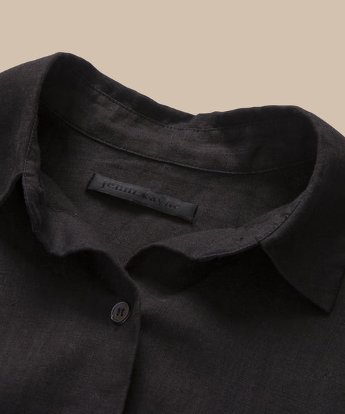 A close-up of the Linen Logan Shirt, a black boxy-fit button-up with a single button visible and an inner collar tag reading jenn kayne, set against a beige background.