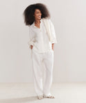 A woman with voluminous curly hair smiles in a loose white button-up, Linen Logan Pant in ivory, open-toe sandals, and a draped sweater—an effortlessly stylish everyday look.