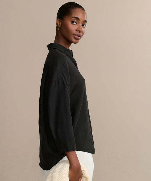A woman with pulled-back hair wears the Linen Logan Shirt in black and white pants, holding a textured beige accessory, against a plain beige background.