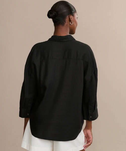 A person with hair in a neat bun, wearing the Linen Logan Shirt styled with rolled sleeves and paired with white shorts, stands facing away against a plain beige background.