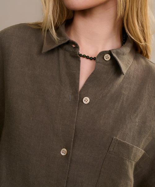 A person with long blonde hair wears the olive-green Linen Logan Shirt—boxy fit, chest pocket—paired with a short black beaded necklace for an effortless style. The face is partially out of frame.