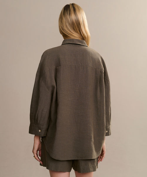 A person with light blonde hair is seen from behind, wearing an oversized, long-sleeve Linen Logan Shirt and matching brown shorts, showcasing effortless style against a plain beige background.