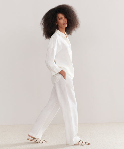 A woman with voluminous curly hair wears a loose white button-up and Linen Logan Pant in ivory, paired with white sandals. She stands against a light background, looking confidently at the camera—perfect for everyday outfitting.