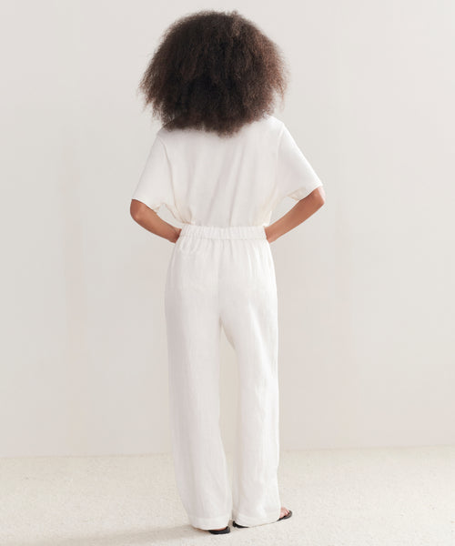 A person with voluminous curly hair stands facing away, wearing a loose white top and Linen Logan Pant—perfect for effortless everyday style. Hands on hips, set against a plain, light-colored background.
