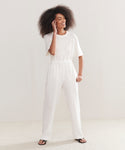 A person with curly hair smiles and touches their face, wearing a loose white t-shirt, the Linen Logan Pant in ivory, and black sandals—an effortlessly chic everyday look set against a plain light backdrop.