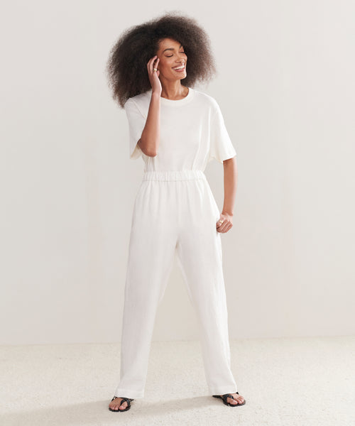 A person with curly hair smiles and touches their face, wearing a loose white t-shirt, the Linen Logan Pant in ivory, and black sandals—an effortlessly chic everyday look set against a plain light backdrop.