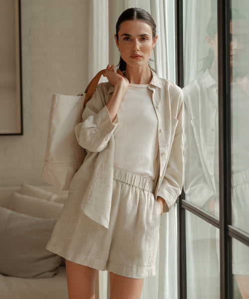 A woman with long dark hair pulled back stands indoors by a window, wearing a light beige 100% linen boxy-fit Linen Logan Shirt, t-shirt, and shorts. She carries a large beige tote bag over her shoulder.