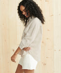 A woman with curly hair, wearing the Linen Logan Shirt in light beige and white shorts, smiles while looking down against a light wooden wall.