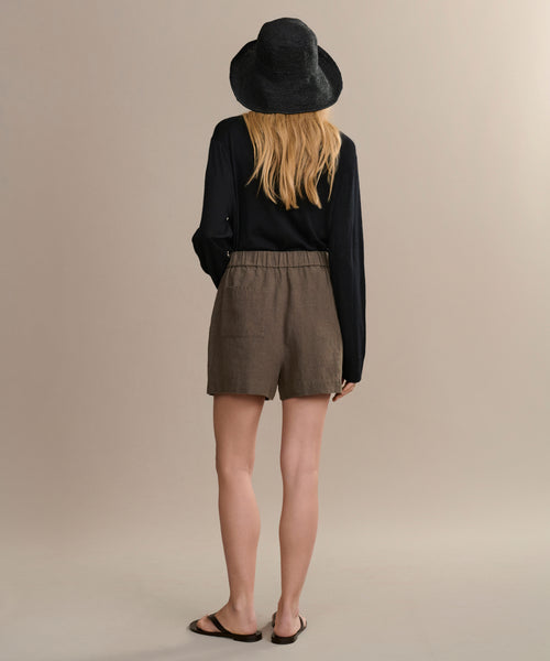 A person with long blonde hair, in a black hat, black long-sleeve top, Linen Logan Short in dark moss, and black sandals stands facing away against a beige background—showcasing the warm-weather style of the Linen Logan Short.