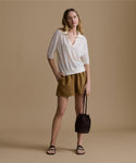 Woman wearing a white shirt and brown shorts, holding a brown bag against a beige background