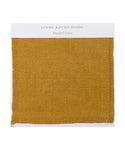 The Fabric Swatch Mustard Linen is a square sample of mustard-colored linen with a visible woven texture and a white label at the top that says “Jenni Kayne Home, Mustard Linen.”.