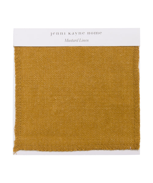 The Fabric Swatch Mustard Linen is a square sample of mustard-colored linen with a visible woven texture and a white label at the top that says “Jenni Kayne Home, Mustard Linen.”.