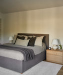 A modern bedroom featuring the Pacific Bed with a gray upholstered headboard, neutral bedding, and throw pillows. A wooden nightstand holds a lamp, candle, and flowers. Light walls and minimal decor create a soothing retreat.